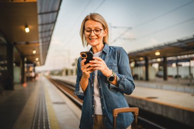 Olgun kadın treni bekle ve tren istasyonunda sıcak içecekle birlikte cep telefonu ve termos kullan.