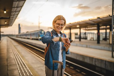 Olgun kadın treni bekle kol saatinde saat kaç, cep telefonu kullan ve tren istasyonunda bavulla yürü.