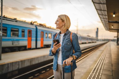 Olgun kadın treni bekle, cep telefonu kullan ve tren istasyonunda bavulla yürü.