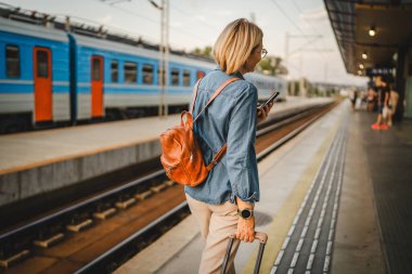 Yetişkin bir kadının arka görüntüsü treni bekle, cep telefonu kullan ve tren istasyonunda bavulla yürü.
