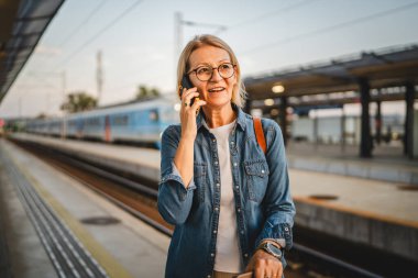 Olgun kadın treni bekle ve beklerken cep telefonuyla konuş.