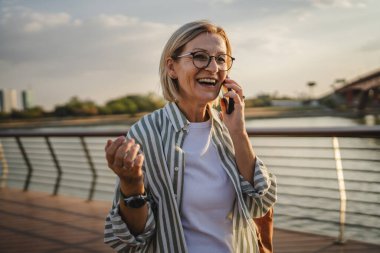Olgun bir kadın cep telefonuyla arıyor ve nehrin önündeki köprüde duruyor.