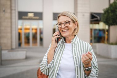 Mutlu olgun bir kadın sokakta yürüyor ve cep telefonuyla konuşuyor.