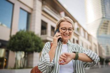 Olgun bir kadın sokakta cep telefonuyla konuşurken kol saatine bakar.