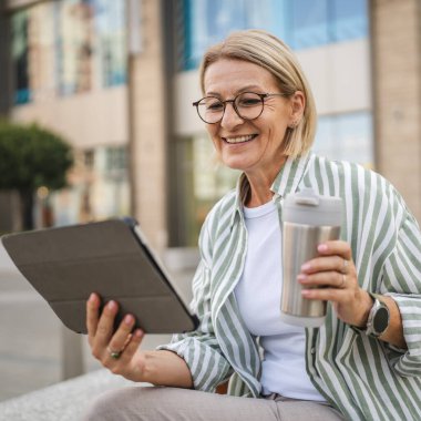 Mutlu olgun bir kadın termosla oturur ve sokakta dijital tablet kullanır.