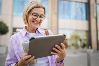 Mutlu olgun bir kadın termosla oturur ve sokakta dijital tablet kullanır.