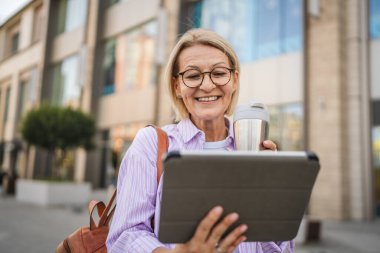 Mutlu olgun bir kadın termosla oturur ve sokakta dijital tablet kullanır.