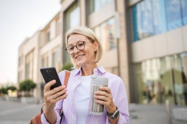 Mutlu olgun bir kadın termosla yürüyor. Sokakta cep telefonu kullanıyor.
