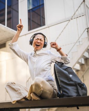 Mature japanese woman listen music on headphones in front the modern buildings clipart