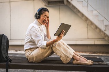 Kulaklıklı olgun Japon kadın, inşaat şirketinin önündeki dijital tablette komik videolar izliyor.