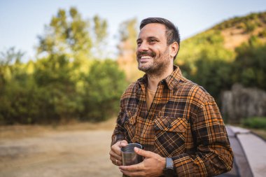 Yetişkin beyaz bir adam kamp gezisinde çadırın önünde sıcak kahve içer.