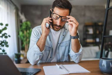 Yetişkin, yorgun, Kafkasyalı adam evde cep telefonuyla konuşuyor.