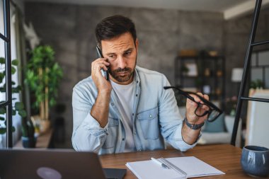Yetişkin, yorgun, Kafkasyalı adam evde cep telefonuyla konuşuyor.