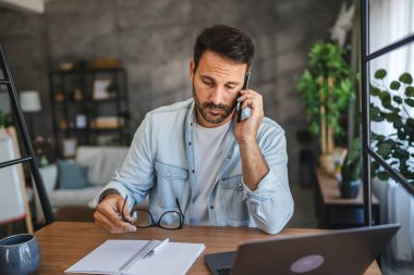 Yetişkin, yorgun, Kafkasyalı adam evde cep telefonuyla konuşuyor.