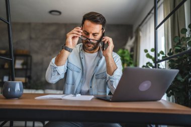 Yetişkin, yorgun, Kafkasyalı adam evde cep telefonuyla konuşuyor.