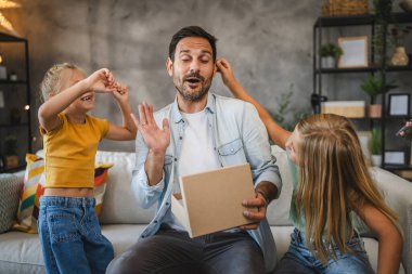 Şaşırmış bir ifadeye sahip neşeli bir baba bir hediye kutusunu tutarken iki kızı da sıcak bir kanepede onu kucaklıyor ve aile eğlencesini kutluyor.