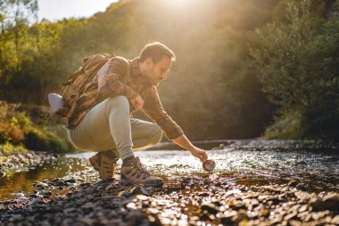 Yetişkin bir adam, yemyeşil bir çayırla çevrili bir bardağı doldurmak için nehrin kenarında çömeliyor. Arka planda bir kamp sandalyesi ve çadır görülür, açık hava macera sahnesi tasvir edilir..