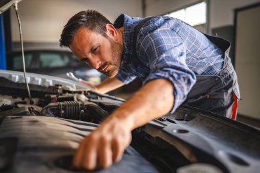 Tulumlu ve ekose gömlekli yetenekli bir araba tamircisi profesyonel bir garaj ortamında çalışırken bir aracın motorunu inceliyor.