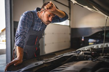 Kareli gömlek ve önlük giymiş yetişkin bir araba tamircisi, iyi ışıklandırılmış, modern bir garaj atölyesinde çeşitli aletlerle özenle çalışırken görülüyor.