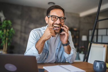 Yetişkin adam evde cep telefonuyla konuşurken gülümseyip eğleniyor.