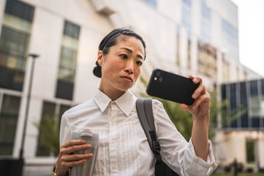 Ciddi yetişkin Japon kadın cep telefonuyla video izleyip termos tutuyor.