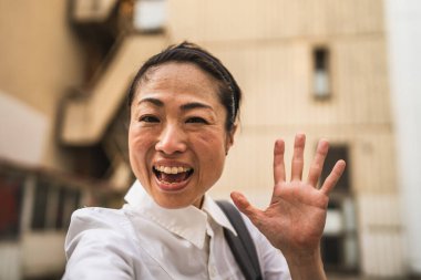 Yetişkin Japon iş kadınının portresi. Sırt çantasıyla modern binaların önünde duruyor. Selamlar.