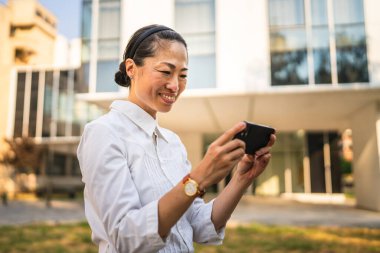 Olgun Japon kadın modern binaların önünde cep telefonuyla video oyunu oynuyor.