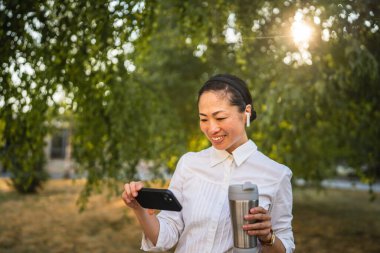Kulaklıklı olgun Japon kadın parkta cep telefonuyla komik videolar izliyor.
