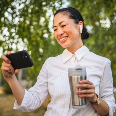 Kulaklıklı olgun Japon kadın parkta cep telefonuyla komik videolar izliyor.