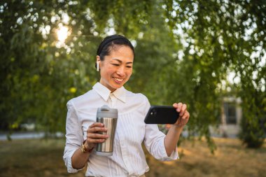 Kulaklıklı olgun Japon kadın parkta cep telefonuyla komik videolar izliyor.