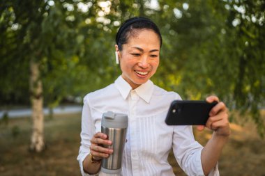 Kulaklıklı olgun Japon kadın parkta cep telefonuyla komik videolar izliyor.