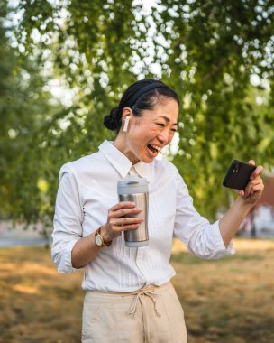Kulaklıklı olgun Japon kadın parkta cep telefonuyla komik videolar izliyor.