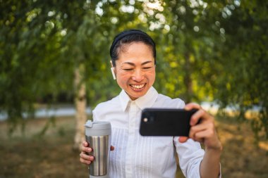 Kulaklıklı olgun Japon kadın parkta cep telefonuyla komik videolar izliyor.