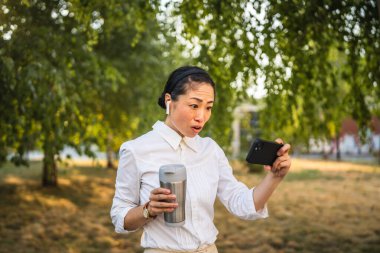 Kulaklıklı olgun Japon kadın parkta cep telefonuyla komik videolar izliyor.