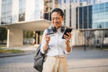 Kulaklık ve sırt çantalı olgun Japon kadın cep telefonundan kredi kartıyla online alışveriş yapıyor.