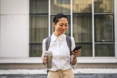 Mutlu olgun Japon iş kadını cep telefonunu açık havada kullan ve termosu tut.