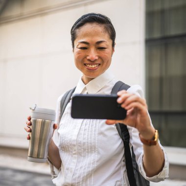 Olgun Japon kadın dışarıda cep telefonuyla komik videolar izleyip termosu tutuyor.