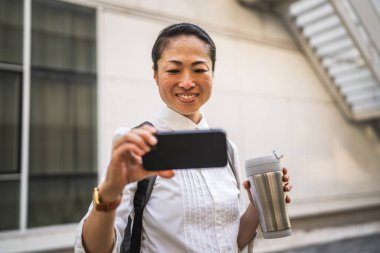 Olgun Japon kadın dışarıda cep telefonuyla komik videolar izleyip termosu tutuyor.