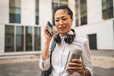 Olgun Japon iş kadını cep telefonuyla konuşur, termosu tutar ve sırt çantası takar.