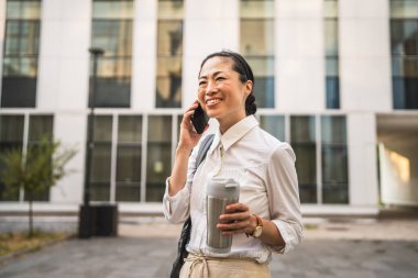 Olgun Japon iş kadını cep telefonuyla konuşur, termosu tutar ve sırt çantası takar.