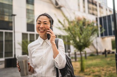 Olgun Japon iş kadını cep telefonuyla konuşur, termosu tutar ve sırt çantası takar.