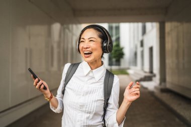 Neşeli Olgun Japon kadın Whit kulaklıklar modern binalar arasında cep telefonundan müzik dinliyor.