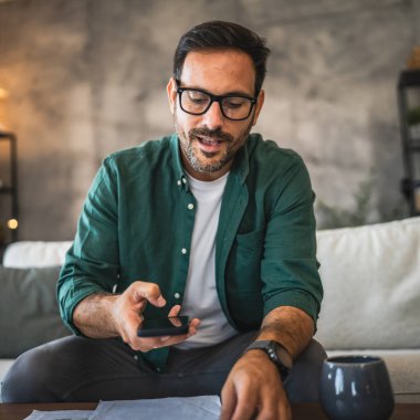 Yetişkin bir adam kanepeye oturur ve cep telefonuna faturalarını öder.