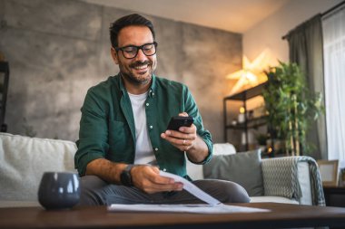 Yetişkin bir adam kanepeye oturur ve cep telefonuna faturalarını öder.