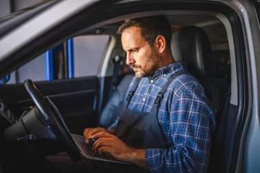 Tulum giymiş odaklanmış bir araba tamircisi teşhis için arabanın içinde dizüstü bilgisayar kullanır. Bu da profesyonel bir garajda modern otomotiv tamir teknolojisi olduğunu gösterir.