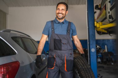 Atölyedeki neşeli araba tamircisinin portresi bir arabanın yanında lastik tutar, tulum ve eldiven giyer. Bu görüntü otomotiv alanında profesyonelliği ve memnuniyeti ifade eder.