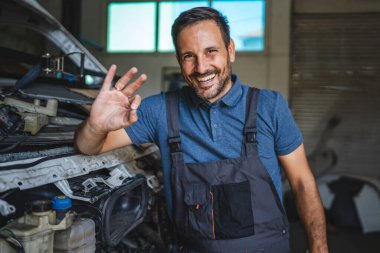Mavi polo ve tulumlu neşeli bir araba tamircisi, bir atölyede bir arabanın yanında durup, olumlu ve profesyonel bir hizmet sunarken 