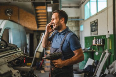 Tulum ve mavi gömlek giyen bir araba tamircisi cep telefonuyla konuşurken matkap tutuyor. Araç gereçleri ve araba parçalarıyla dolu bir oto tamirhanesinde duruyor.