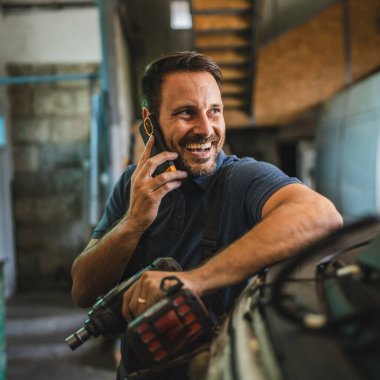 Tulum ve mavi gömlek giyen bir araba tamircisi cep telefonuyla konuşurken matkap tutuyor. Araç gereçleri ve araba parçalarıyla dolu bir oto tamirhanesinde duruyor.