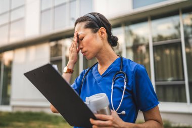 Endişeli yetişkin Japon doktor elinde dosya ve termosla binanın önünde duruyor.